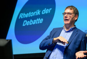 Das Bild zeigt Prof. Olaf Kramer während eines Vortrags. Im Hintergrund ist eine Powerpoint Präsentation zu sehen.
