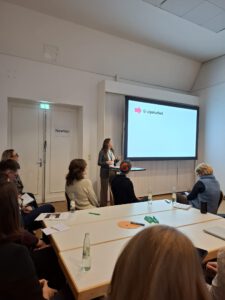 Das Bild zeigt Selina Bernarding im Workshop. Sie steht vor einer Leinwand und doziert. Im Vordergrund sind von hinten fortografiert Menschen an einem Tisch zu sehen, die Selina Bernarding zuhören.